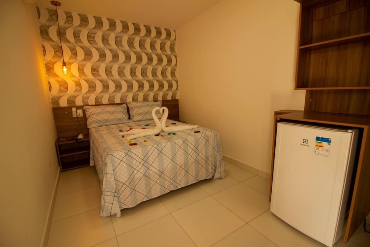 Un dormitori amb un llit, una tauleta de nit, una làmpada penjada a la paret, un frigor ífic i una paret amb un disseny decoratiu.