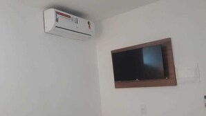 A white air conditioning unit mounted on the wall, a flat-screen TV with a wooden frame, and a power outlet below the TV. - Pousada Aliança (Ipojuca)