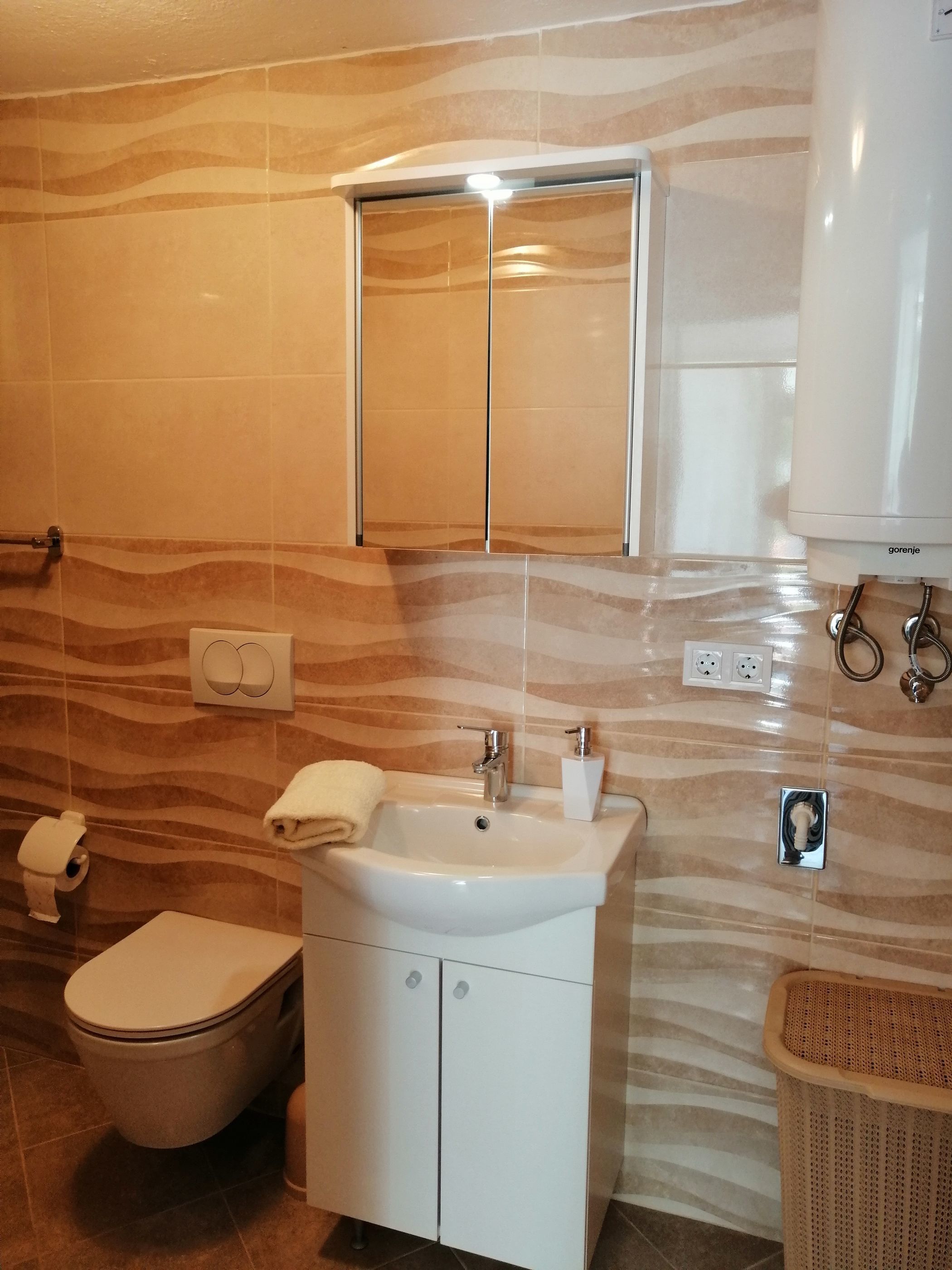 A bathroom with a toilet, sink, and mirror. There is a towel hanging on the wall and a trash bin.