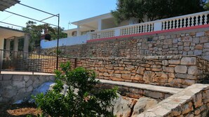 Un mur de pedra amb una tanca de metall, un arbre petit i un edifici amb un balcó al fons.