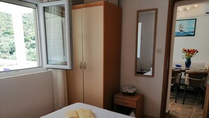 Habitació d'hotel amb un llit, un armari, una taula petita amb un vas de flors i una zona de menjador visible a través d'una porta oberta.