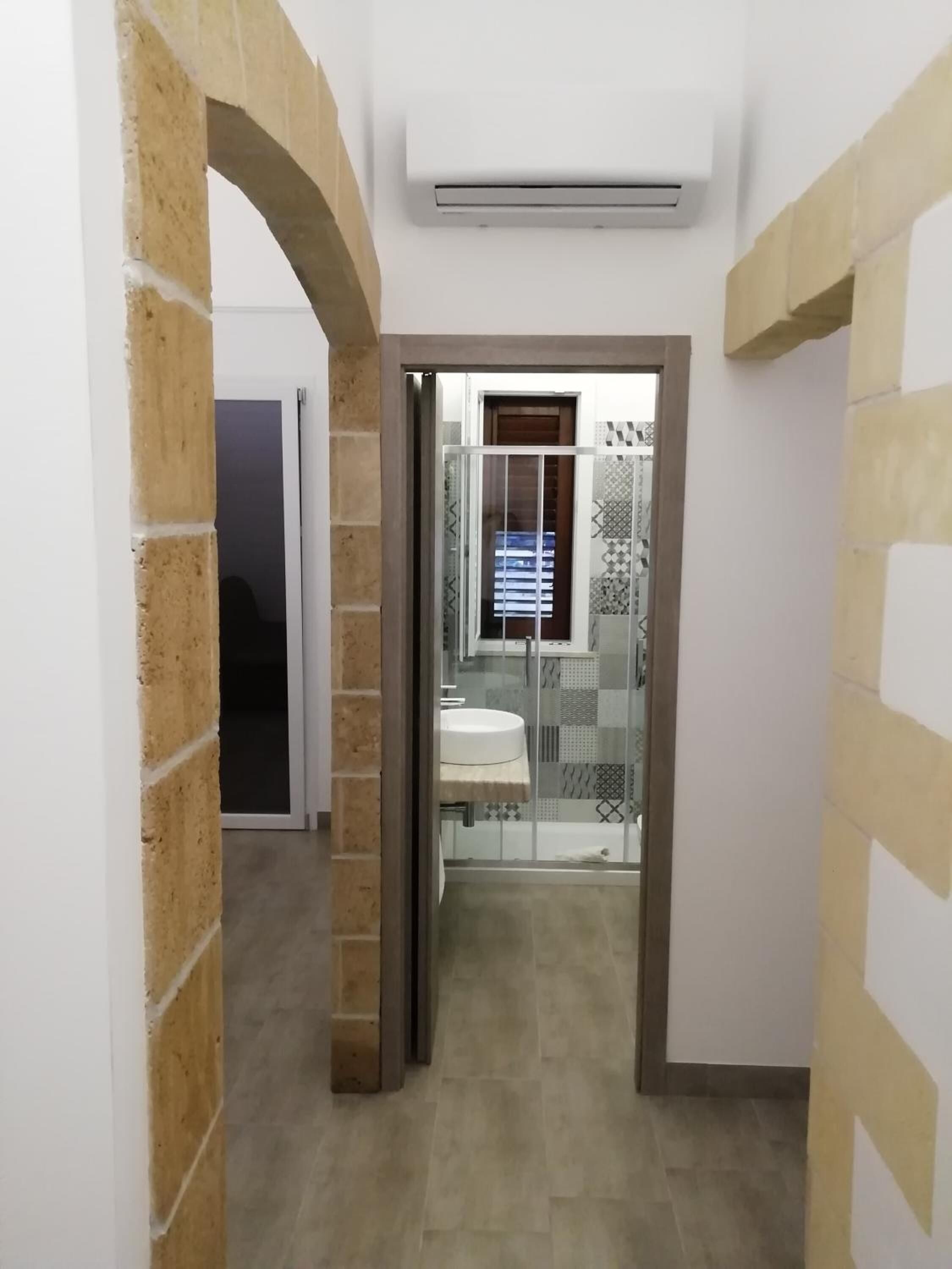 Un couloir étroit doté d’un sol carrelé, une porte donnant sur une salle de bain avec un lavabo et des carreaux décoratifs, et un appareil de climatisation fixé au mur.
