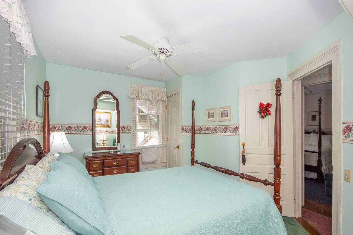 A bedroom with a four-poster bed, a dresser, a mirror, a ceiling fan, and a window with a view of a staircase.