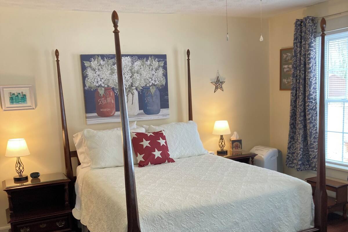 A four-poster bed with a white bedspread and red pillows, a nightstand with a lamp, a ceiling fan, and a window with curtains.