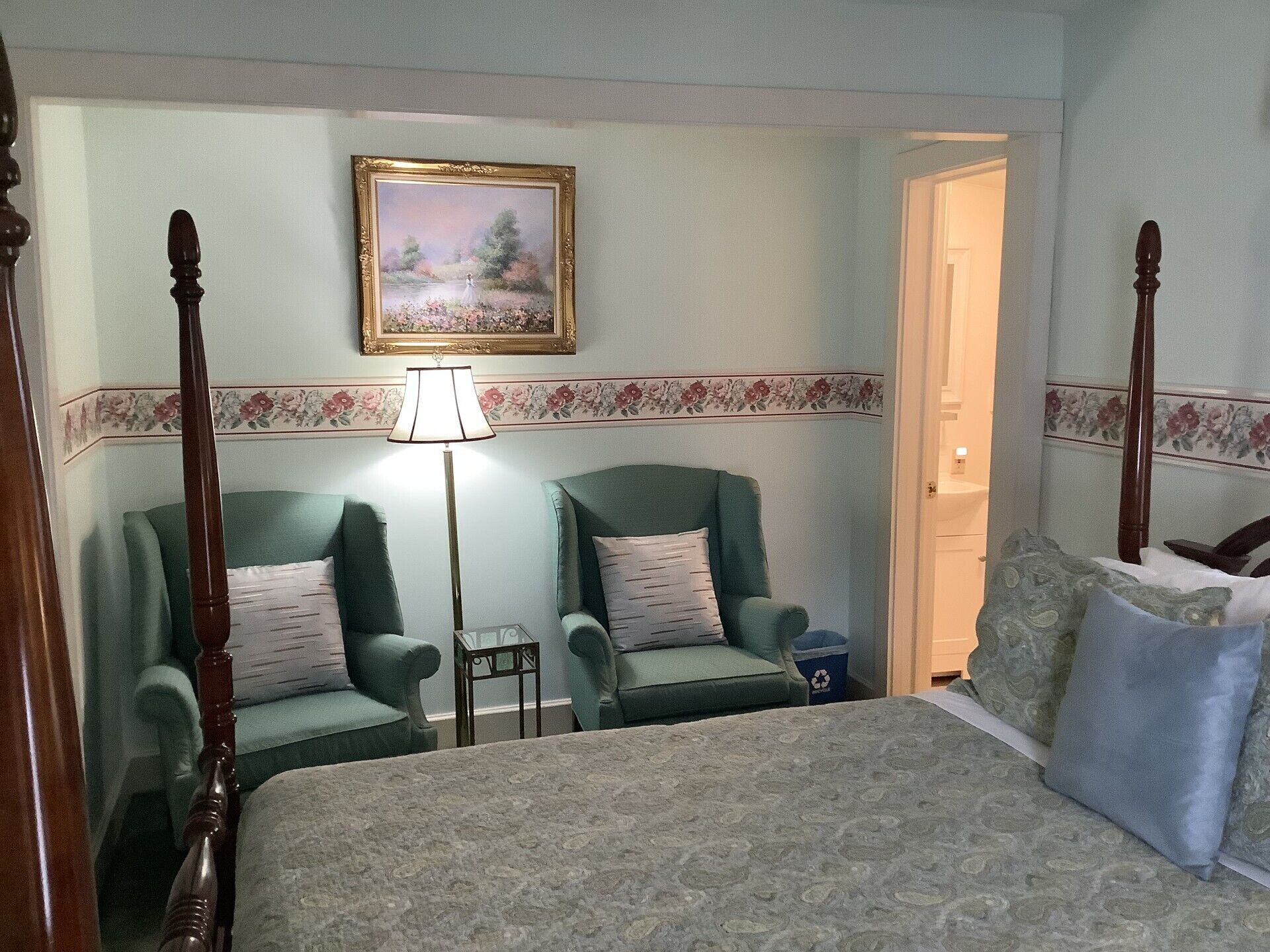 Quarto com cama de dossel, dois poltronas verdes, luminária de chão e um quadro na parede.