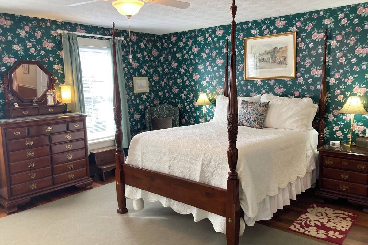 A four-poster bed in a room with floral wallpaper, a ceiling fan, and a large mirror.