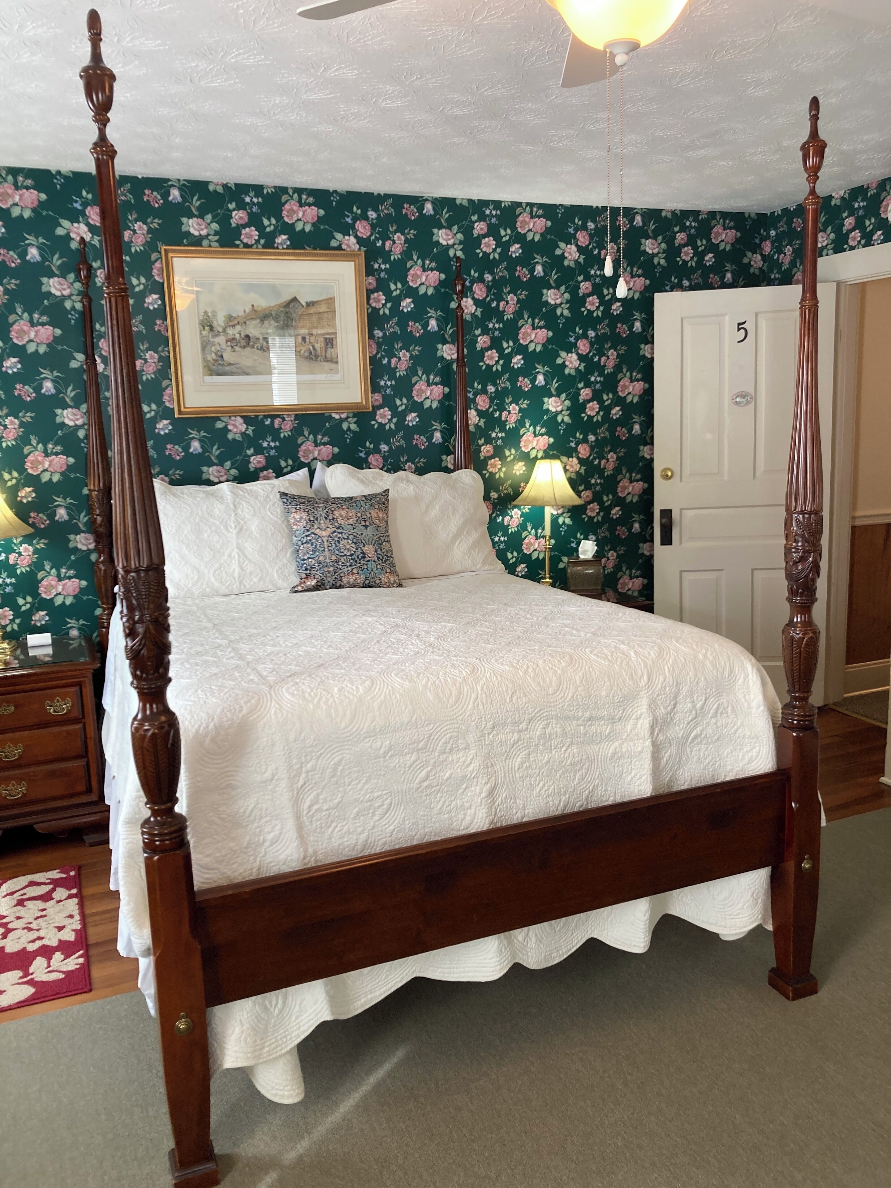 A four-poster bed with a white quilt, a floral wallpaper background, and a framed picture above it.