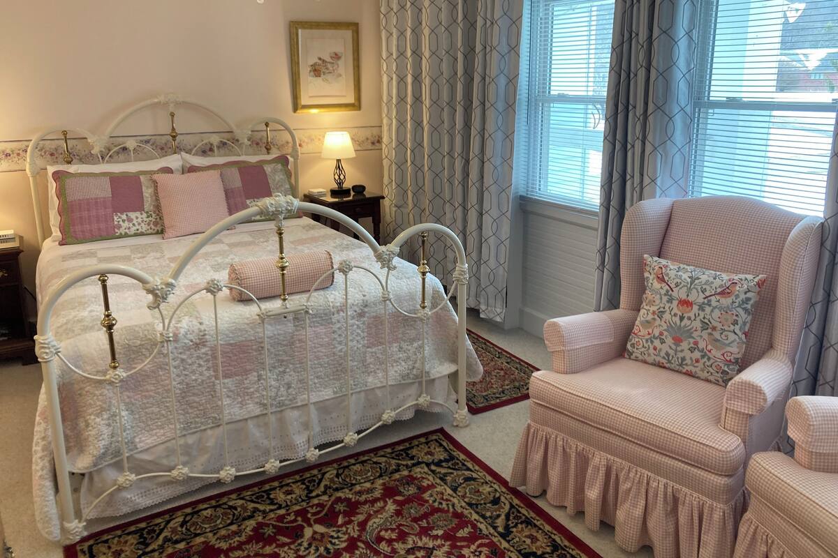A bedroom with a white metal bed frame, a checkered armchair, a patterned rug, and a window with curtains.