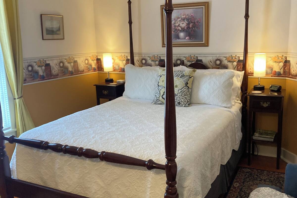 A four-poster bed with a canopy, two bedside tables with lamps, a painting on the wall, and a patterned rug.