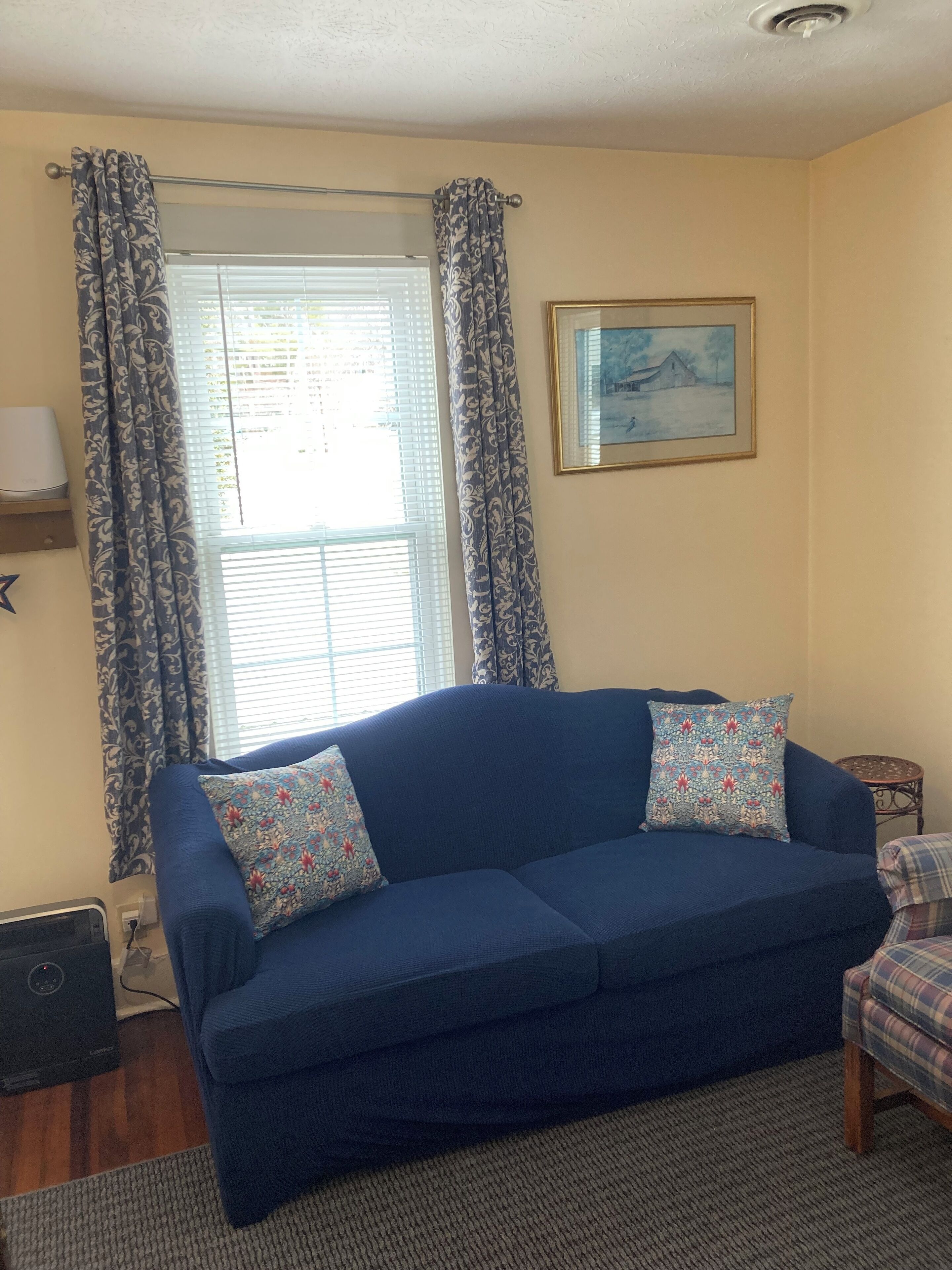 Un salón con un sofá azul, cortinas estampadas, un cuadro en la pared y una pequeña mesa con una silla.