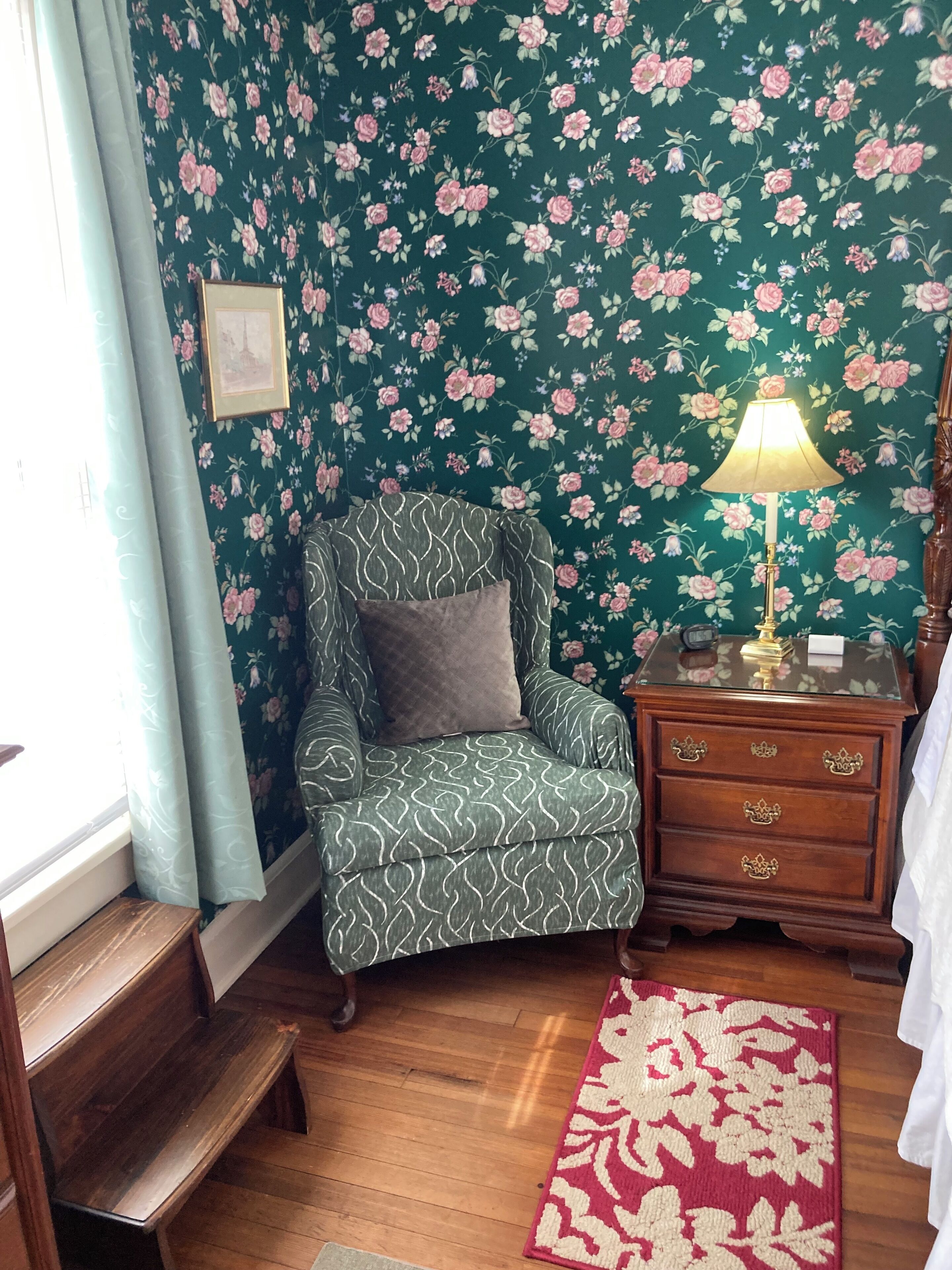 Une pièce avec un papier peint à motifs floraux, un fauteuil vert à motifs, une table de chevet en bois avec des tiroirs, un tapis rouge et blanc, et une fenêtre avec des rideaux.