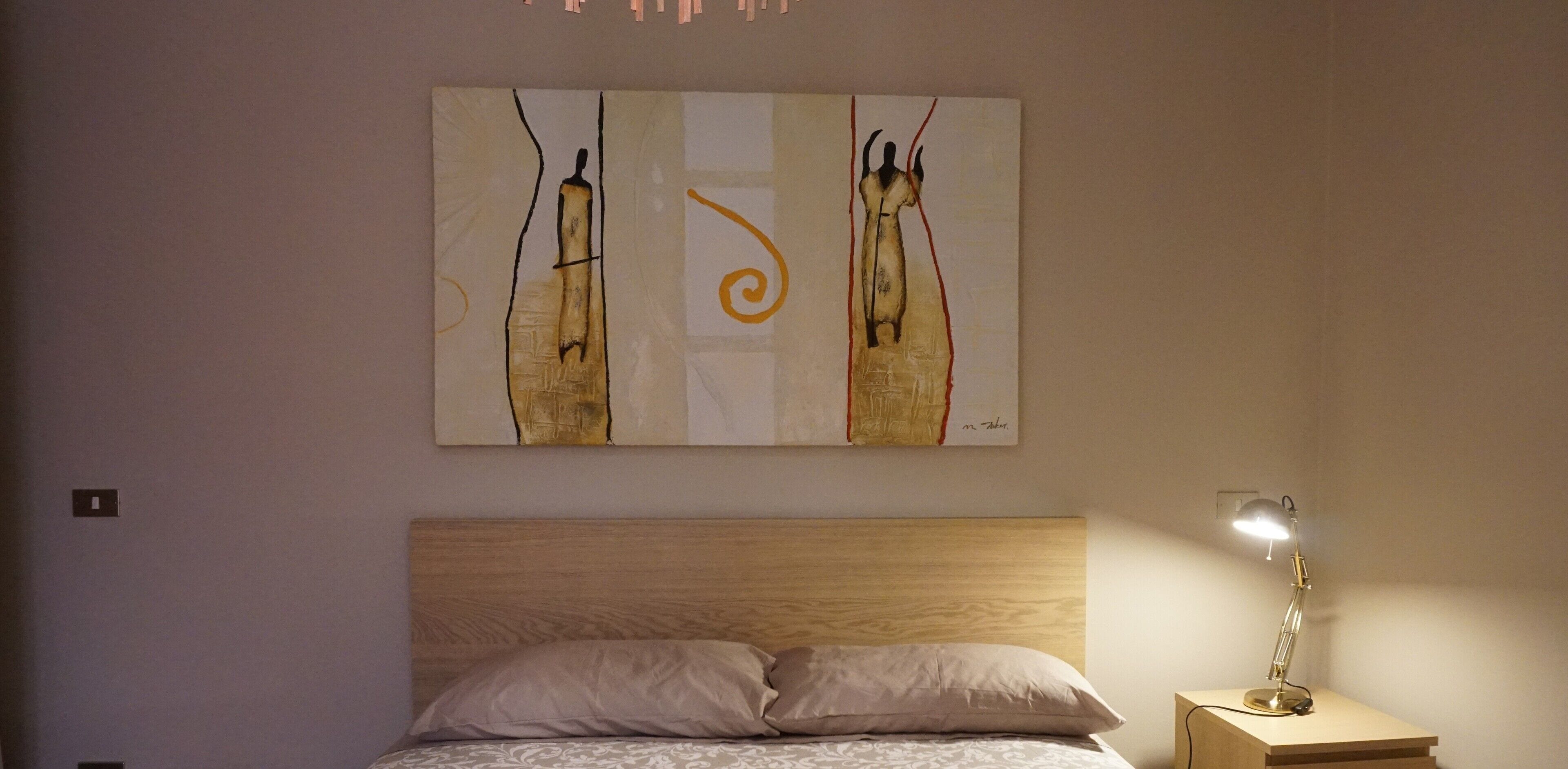 Un dormitorio con cabecera de madera, cama con colcha estampada y mesita de noche con lámpara. Sobre la cama, un cuadro abstracto de gran tamaño.
