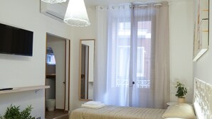 Een slaapkamer met een bed, een aan de muur bevestigde televisie, een nachtkastje met een plant en een raam met dunne gordijnen.