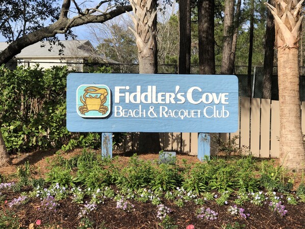 Un panneau indiquant l’entrée de la plage et du club de tennis de Fiddler’s Cove, entouré de verdure et d’arbres.