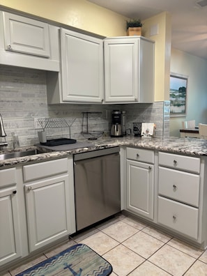 Una cocina con gabinetes blancos, lavavajillas, cafetera y salpicadero de azulejos.