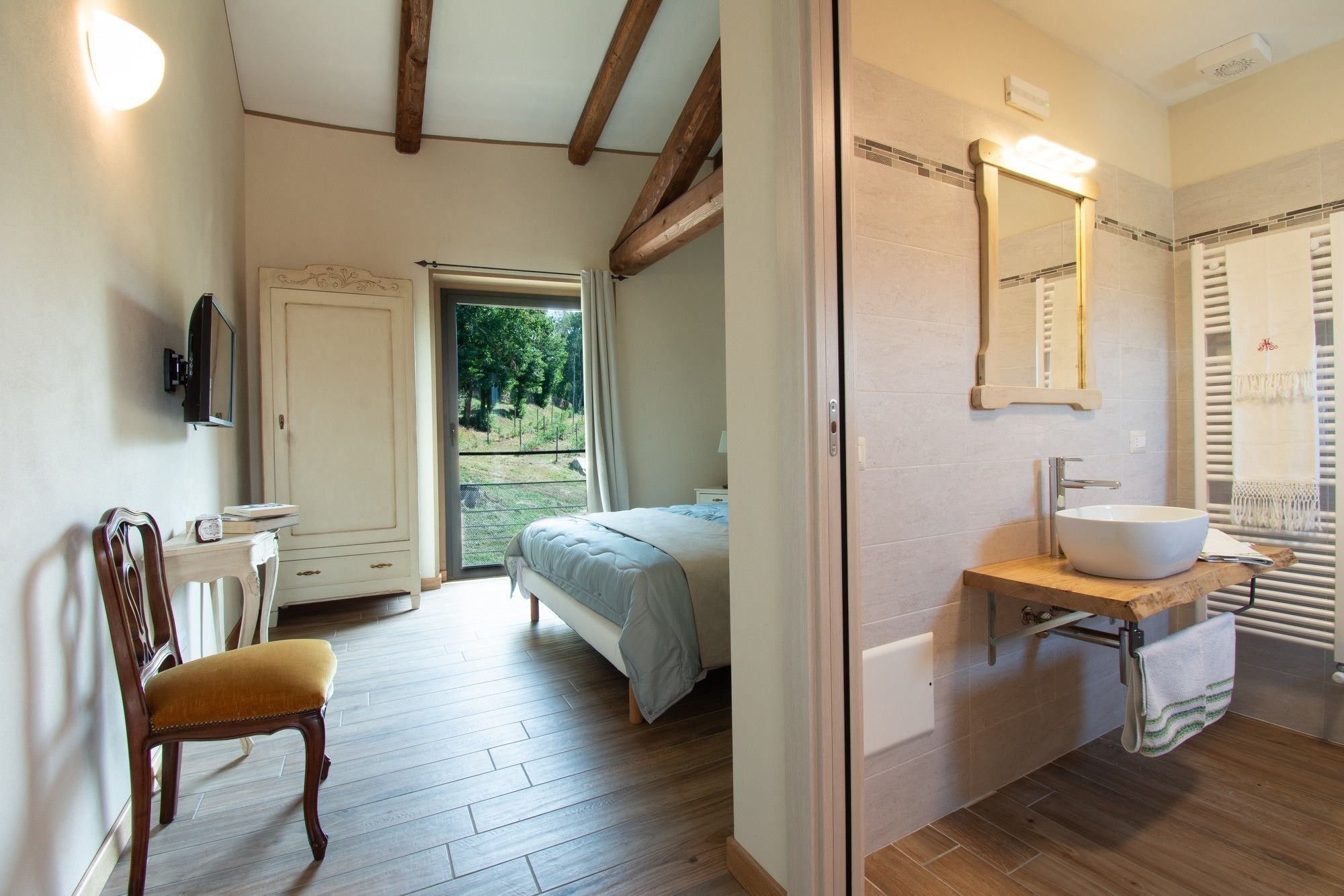 A bedroom with a bed, a chair, a desk, a TV, and a bathroom with a sink and mirror.