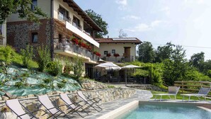 A pool area with lounge chairs and umbrellas, a stone wall, and a building with balconies and red flowers. - Cascina Montiglio (Quagliuzzo)