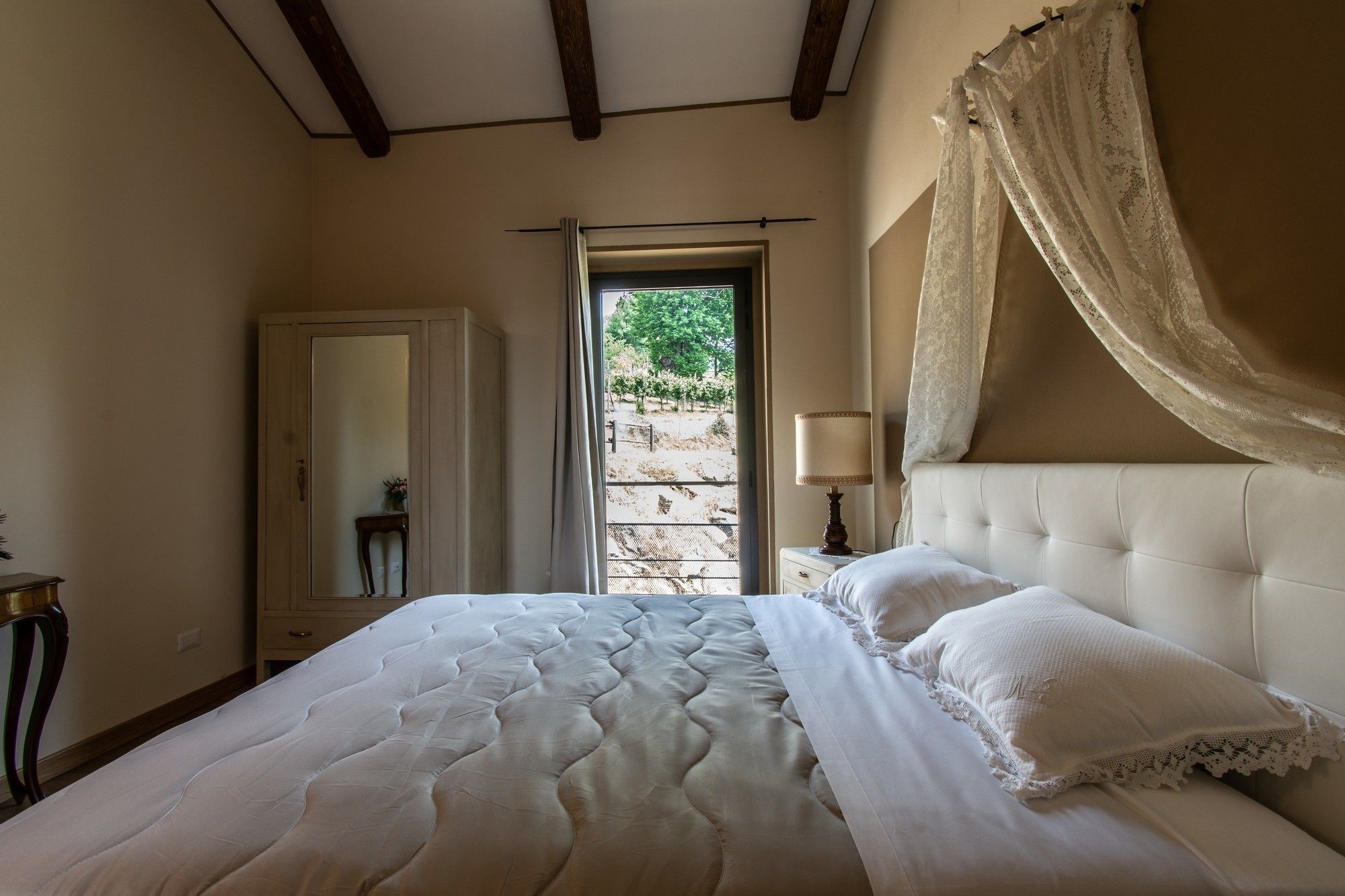 Un dormitorio con una cama grande, una mesita de noche con lámpara, un espejo y una ventana con vistas a la vegetación.