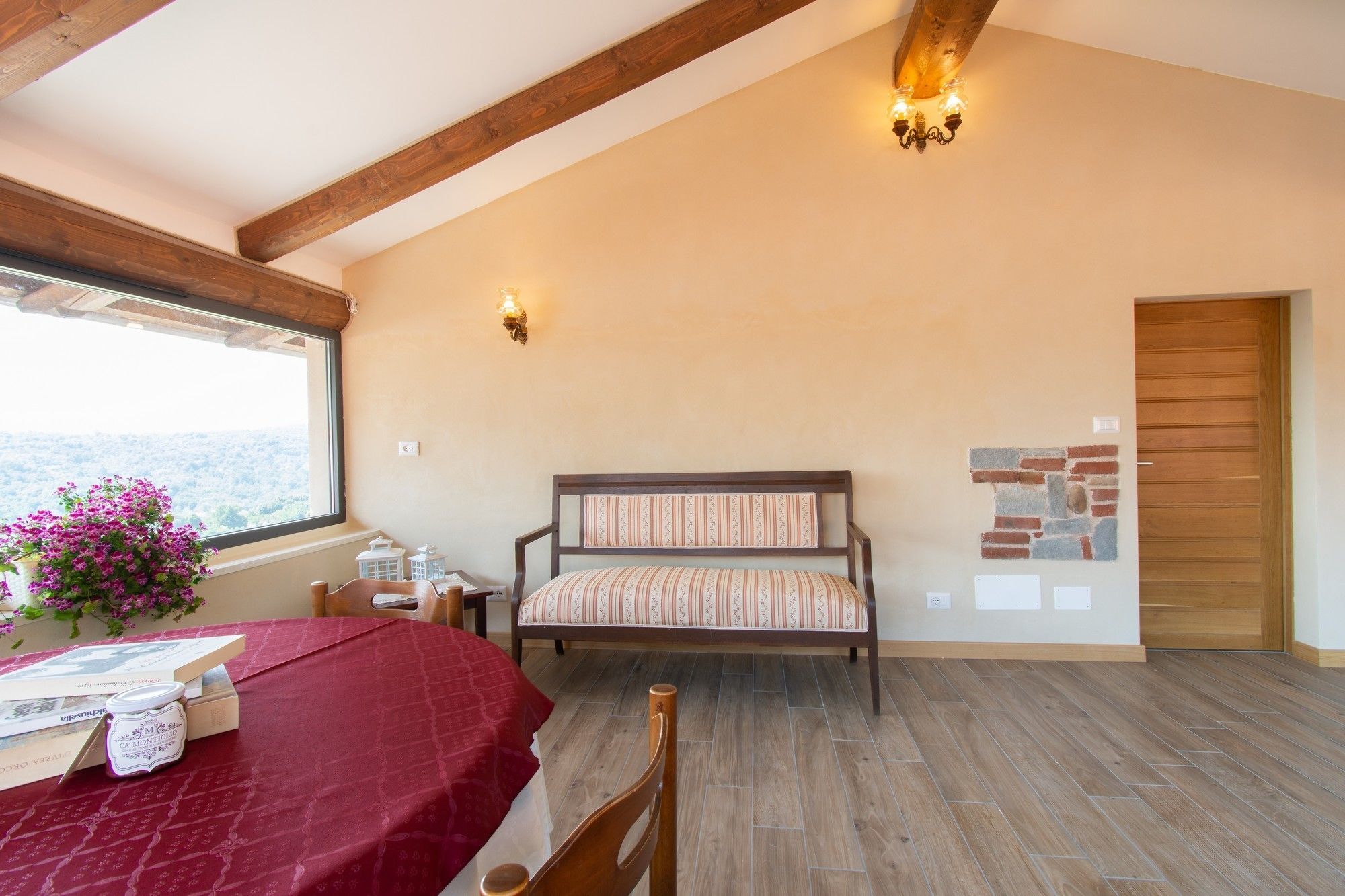Una habitación acogedora con techo de madera, una cama con ropa de cama a rayas, una mesa de madera con mantel rojo y una ventana con vistas.