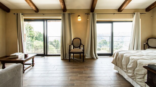 A room with wooden beams, two large windows with curtains, a bed, a chair, and a small table. - Cascina Montiglio (Quagliuzzo)