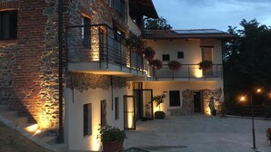 A two-story building with stone and brick walls, a balcony with hanging flower baskets, and a well-lit entrance. - Cascina Montiglio (Quagliuzzo)