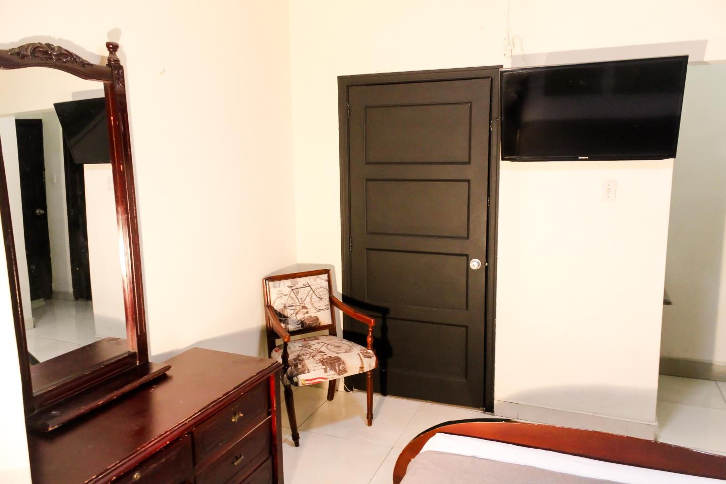 Une chambre avec une commode en bois, une chaise, une porte sombre et un téléviseur à écran plat fixé au mur.