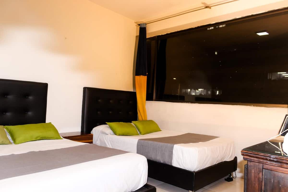 Habitación de hotel con dos camas individuales, cada una con colcha gris y blanca y almohadas verdes. Cuenta con un ventanal grande con cortinas negras, una mesita de noche de madera y una pequeña mesa con lámpara.