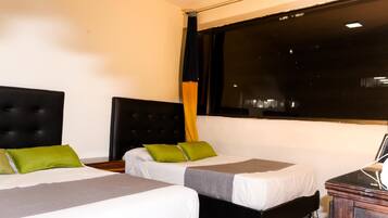 A hotel room with two single beds, each with a grey and white bedspread, and green pillows. There is a large window with black curtains, a wooden nightstand, and a small table with a lamp.