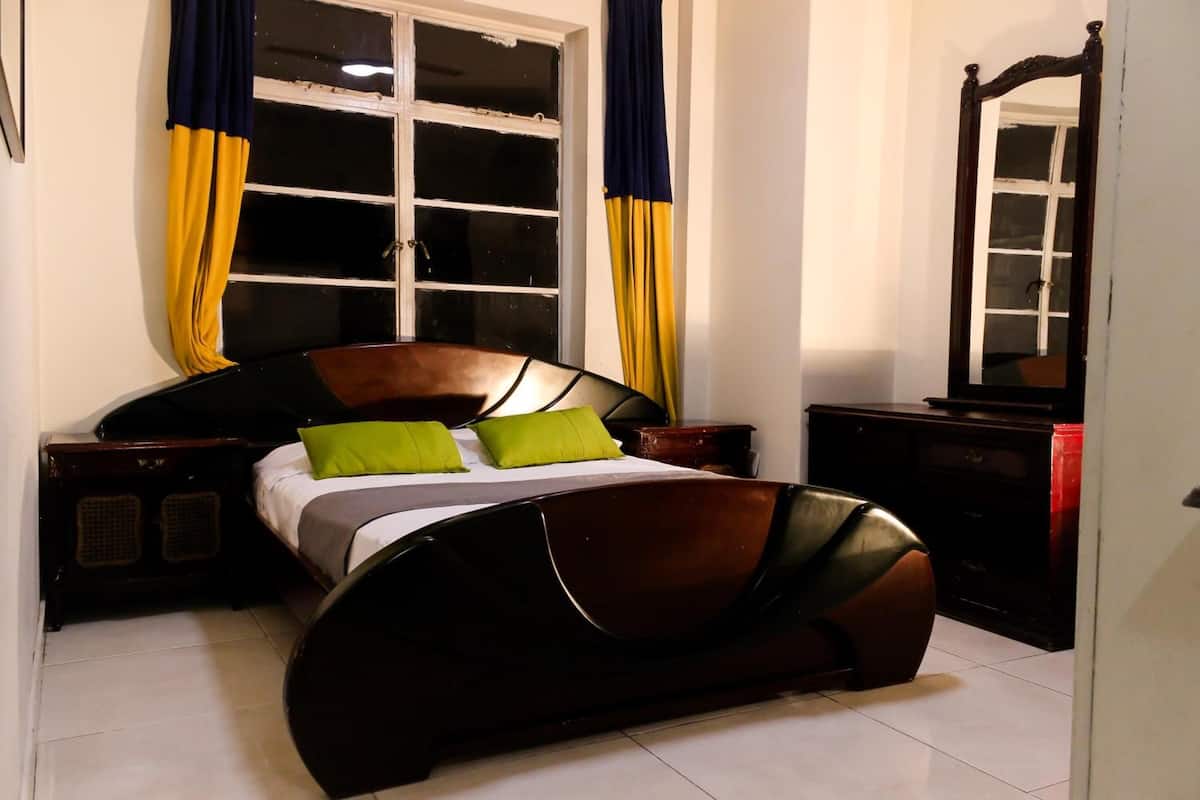 Un dormitorio con cama de madera, almohadas verdes, una mesita de noche, un aparador y una ventana con cortinas.