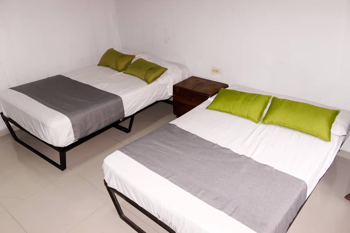 Una habitación con dos camas individuales, cada una con una manta gris y almohadas verdes. Hay una mesita de noche de madera entre las camas.