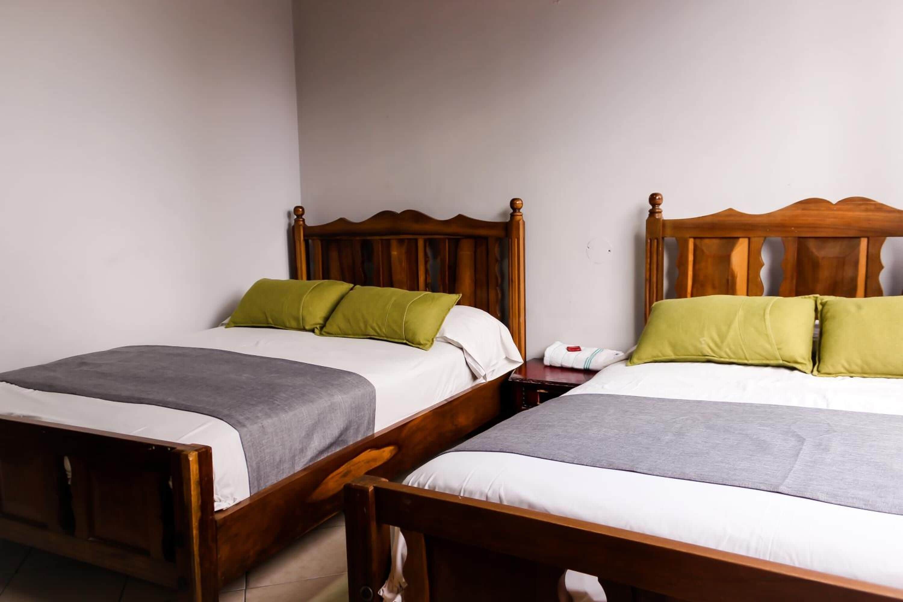 Dos camas de madera con ropa de cama verde y blanca, una mesita de noche con una lámpara y una pared blanca.