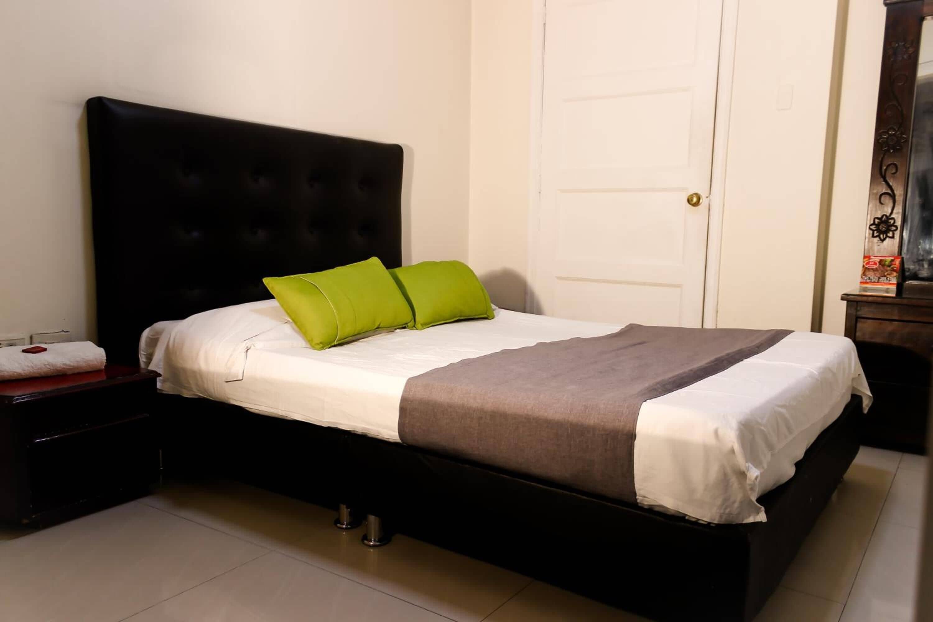 Een netjes opgemaakt bed met een zwart hoofdbord, witte beddengoed en twee groene kussens.