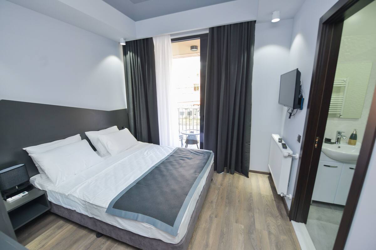 A modern hotel room with a large bed, a bedside table, a TV mounted on the wall, and a bathroom with a sink and mirror.