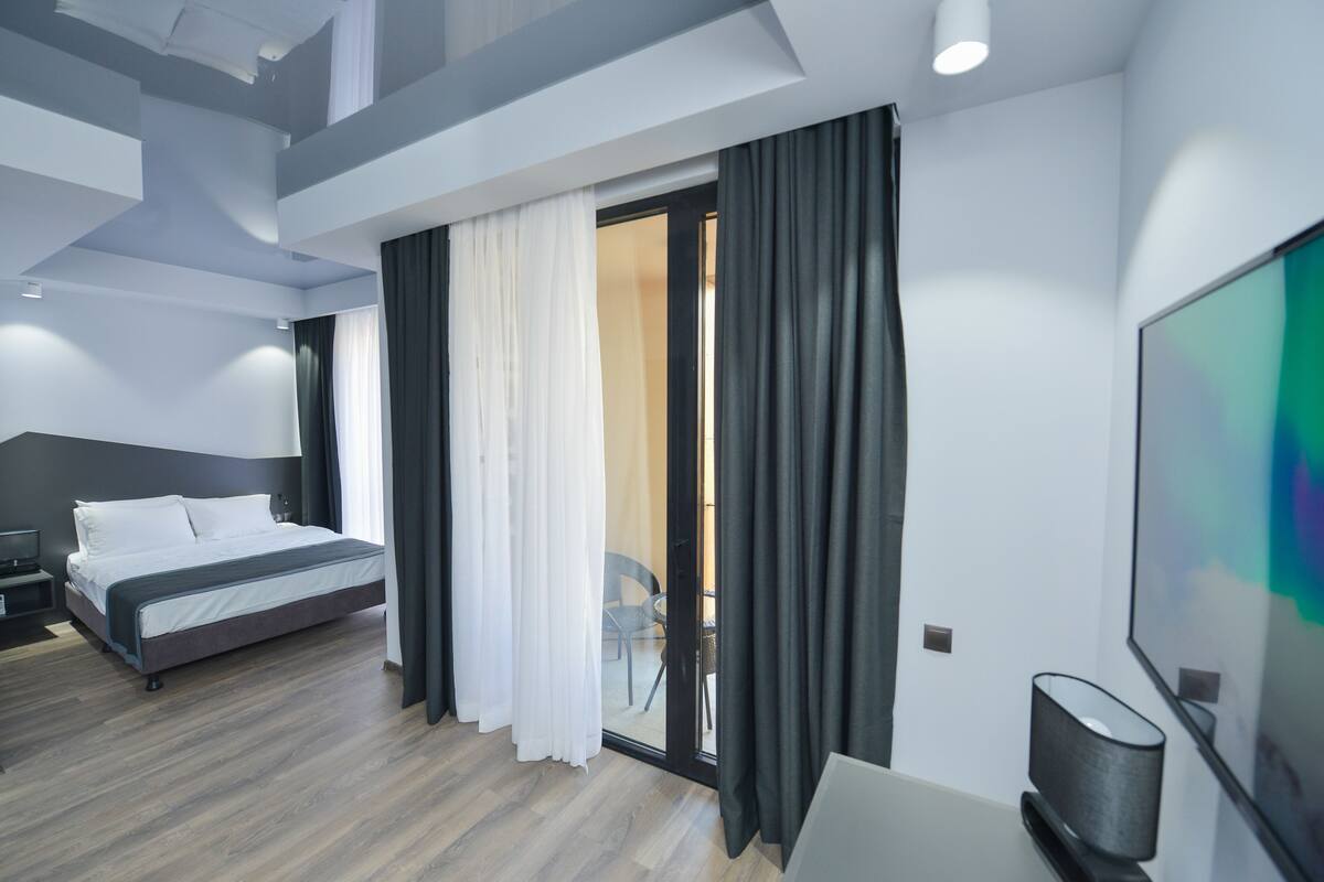 A modern hotel room with a large bed, a desk with a lamp, and a sliding glass door leading to a balcony.