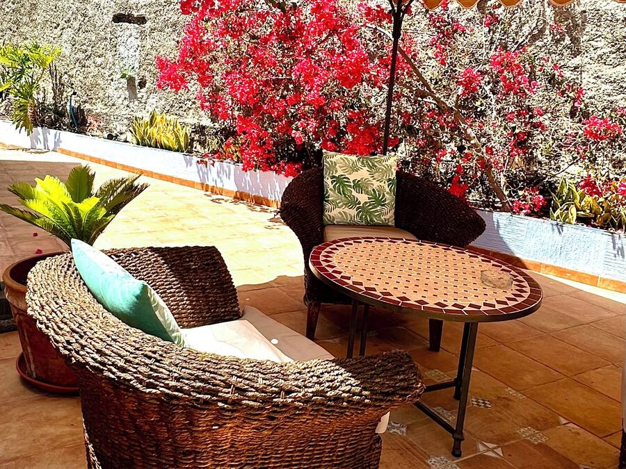 An outdoor seating area with wicker furniture and a large umbrella, surrounded by a garden with red flowers and various plants.