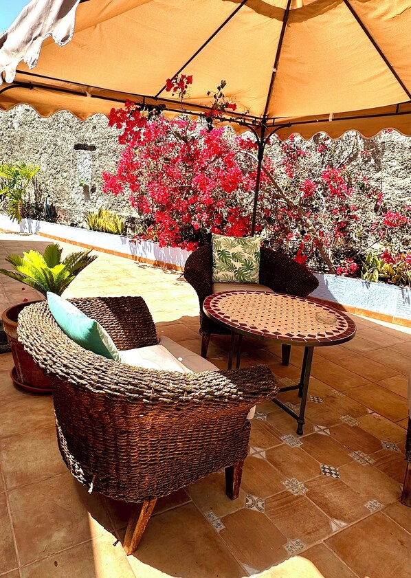 An outdoor seating area with wicker furniture and a large umbrella, surrounded by a garden with red flowers and various plants.