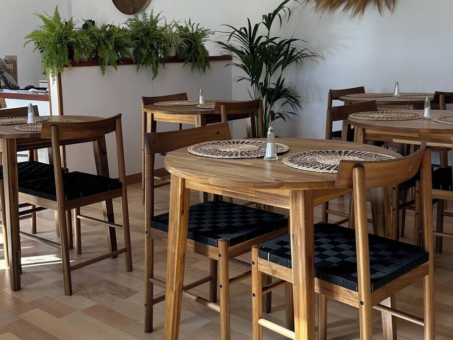 Un comedor con mesas y sillas de madera, un tapiz tejido grande y varias plantas en macetas.