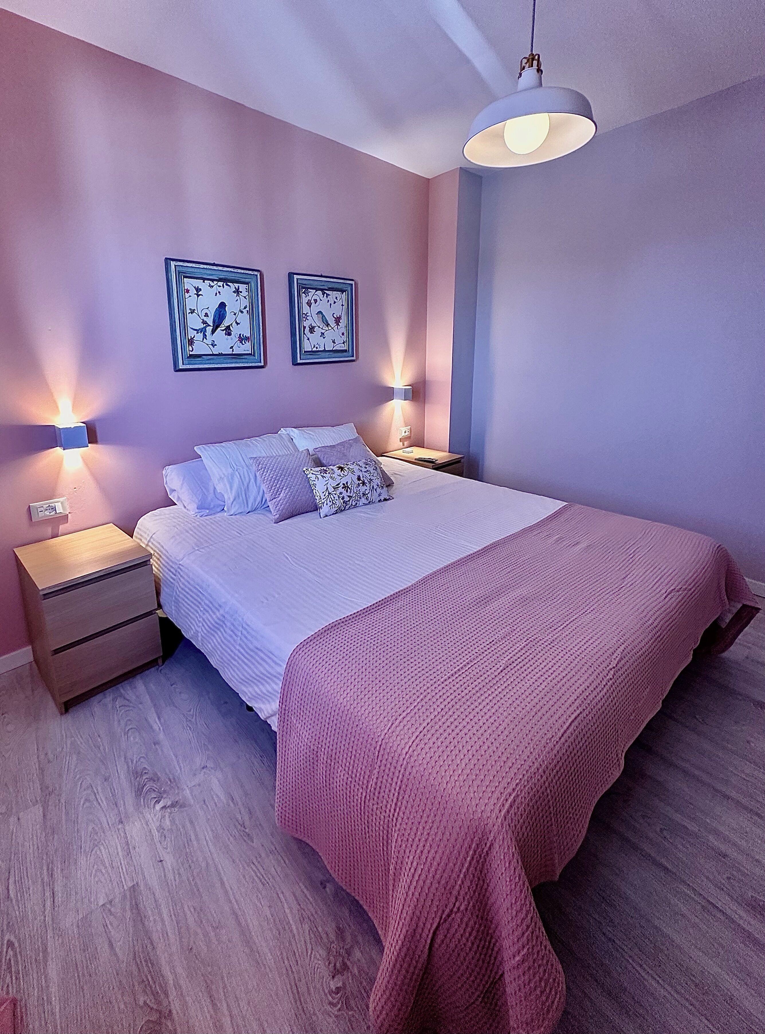 Un dormitorio con una cama, dos lámparas de pared, una mesita de noche y dos cuadros enmarcados.