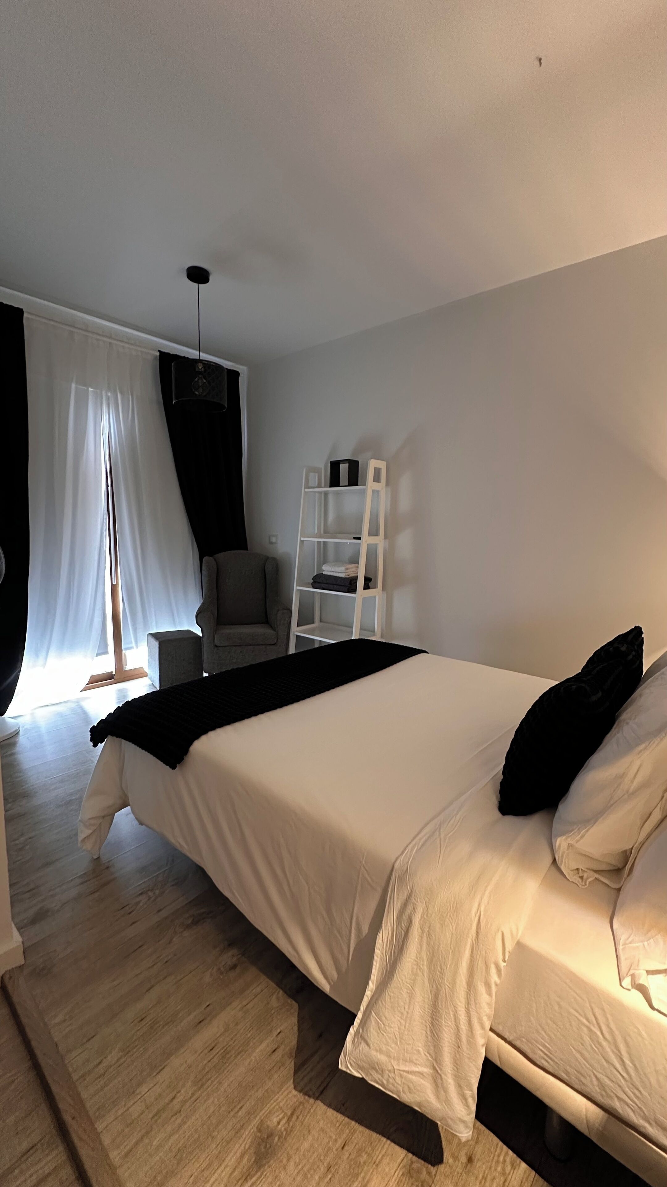 A modern bedroom with a large bed, a chair, a ladder shelf, and sheer curtains.