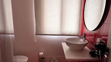 A bathroom with a toilet, a sink with a basin, a mirror, a window with blinds, and a red tiled floor.