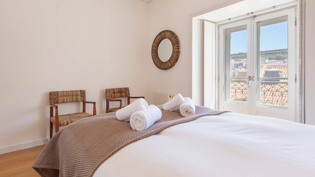 A hotel room with a large bed, two chairs, a round mirror, and a window with a city view. - Downtown Apartment Santa Justa (Lisboa)