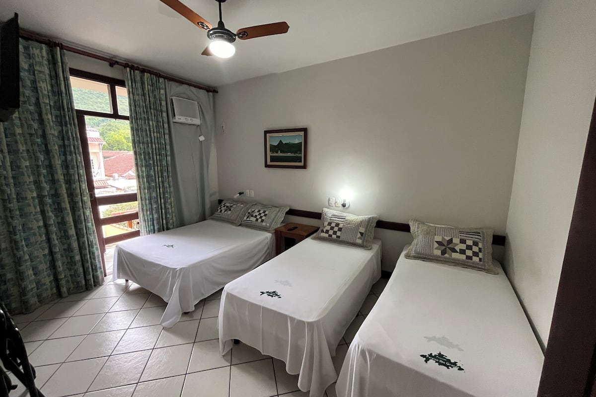 Une chambre d’hôtel avec deux lits simples, un ventilateur de plafond, une fenêtre avec des rideaux et une petite table avec une lampe.