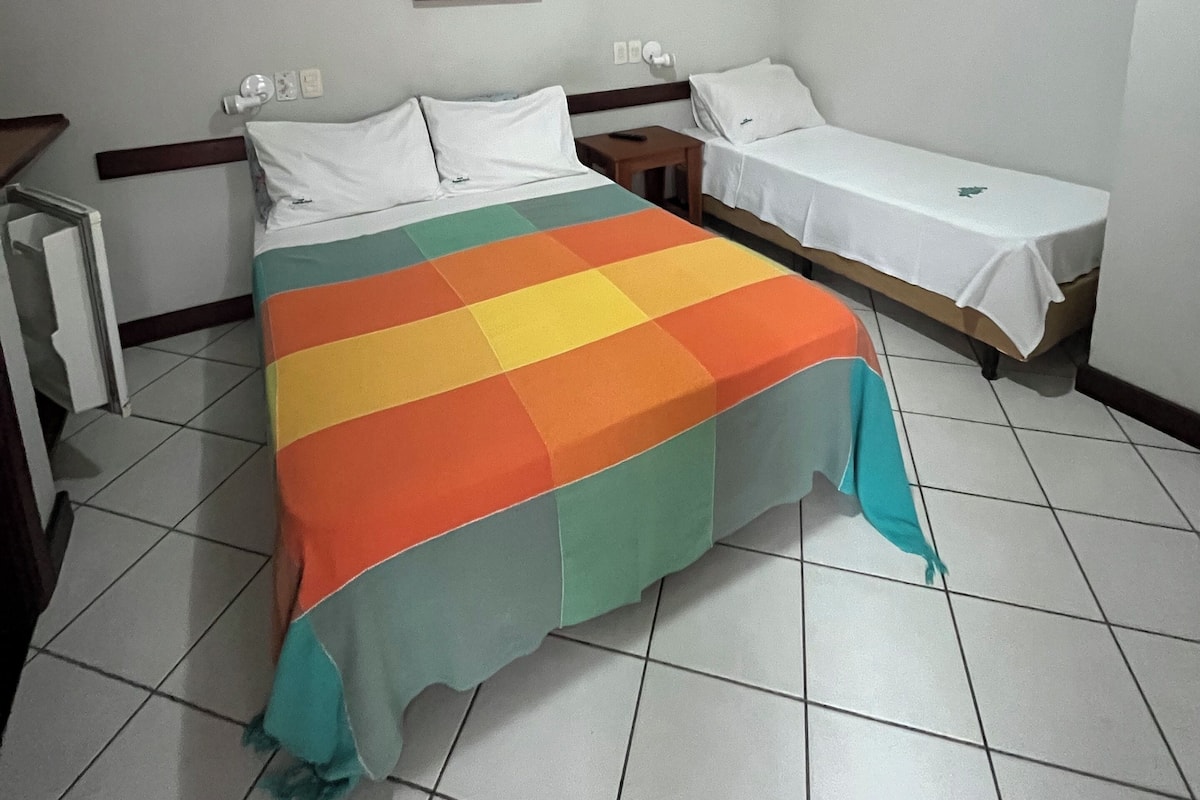 Une chambre d’hôtel avec un lit, une petite table, une chaise, un ventilateur de plafond et un tableau accroché au mur.