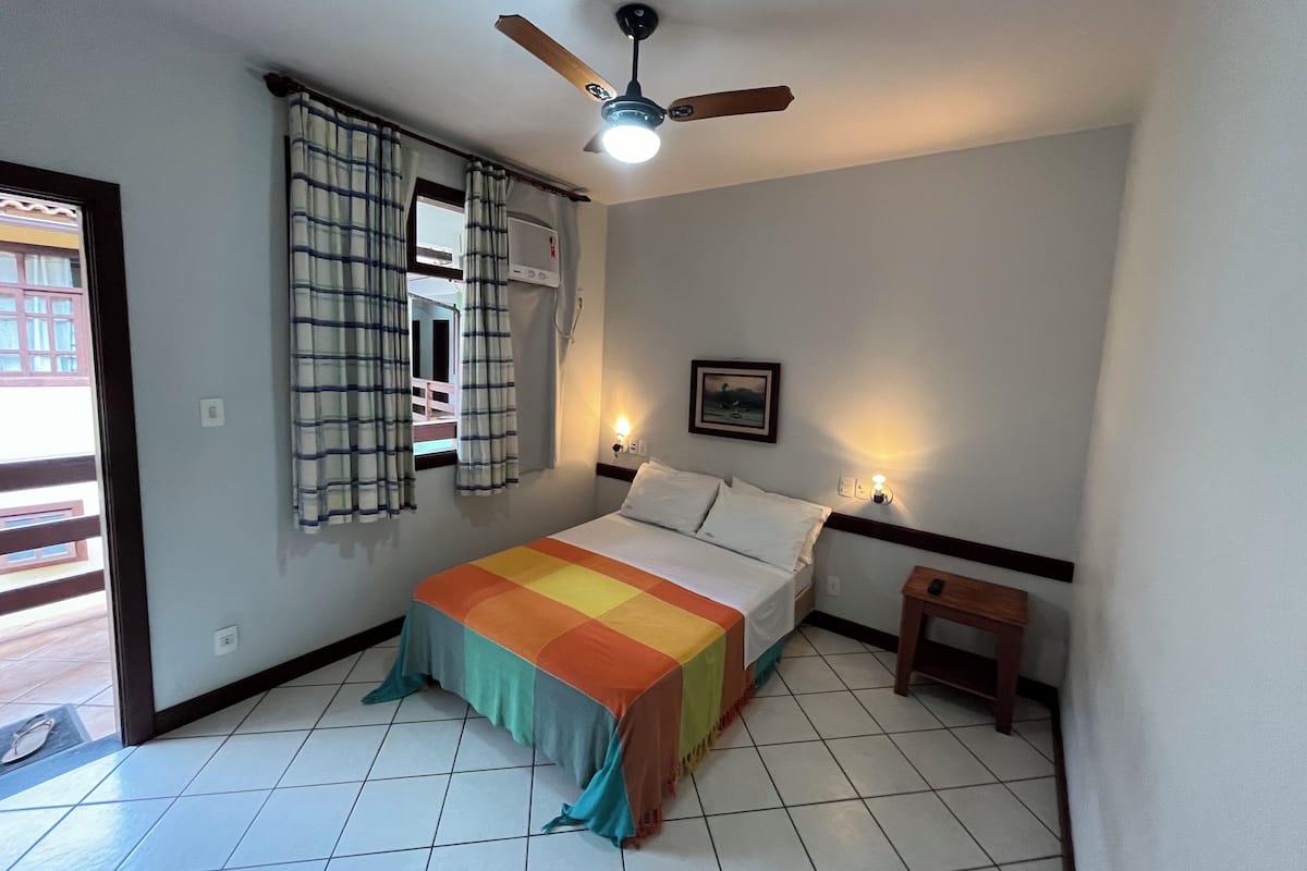 Une chambre à coucher comprenant un lit, un ventilateur de plafond, une fenêtre avec des rideaux, une table de chevet et une lampe fixée au mur.