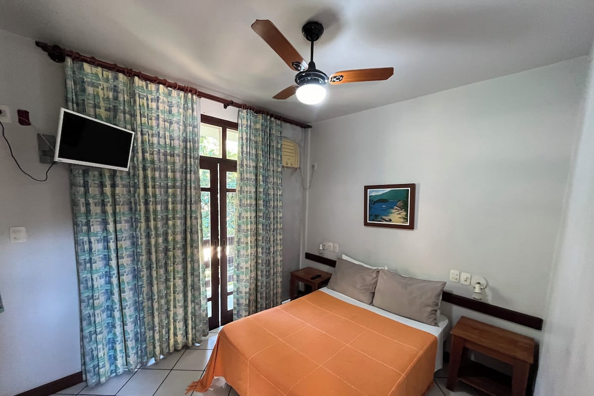 Une chambre d’hôtel avec un lit, un ventilateur de plafond, un téléviseur fixé au mur et un tableau accroché au mur.