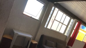 Ein Zimmer mit einem gemusterten Teppich, einem weißen Tisch mit zwei Hockern, einer Couch und großen Fenstern mit Vorhängen.
