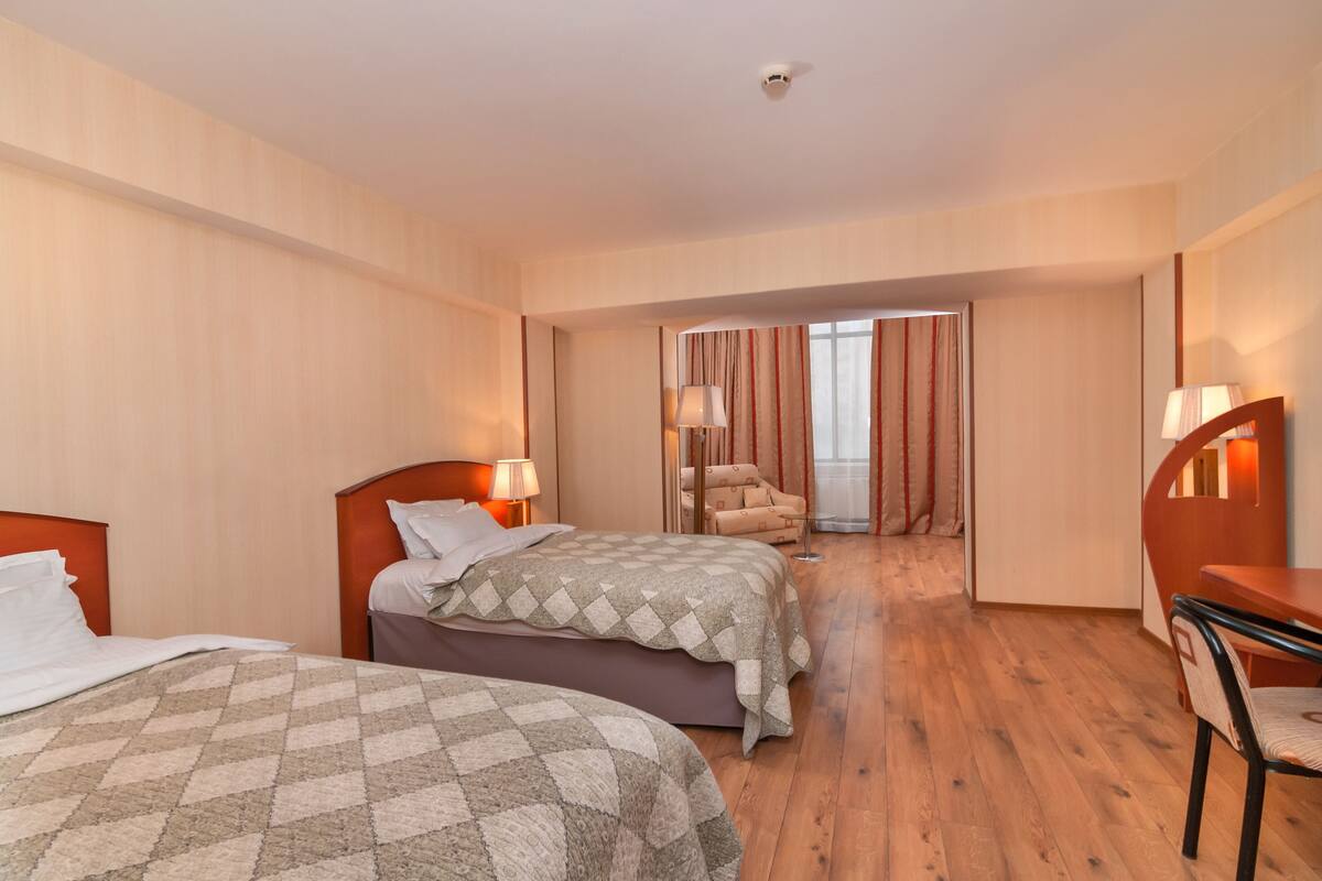 A hotel room with two beds, a desk, a chair, and a view of the outside through a door.