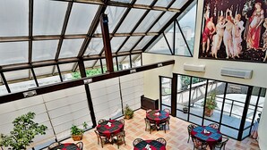 Un espacio interior amplio con techo alto y ventanas grandes, con varias mesas con manteles a cuadros rojos y negros y sillas, y un mural en la pared.