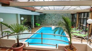 Piscina interior con techo de vidrio, rodeada de plantas en macetas y sillones de descanso.