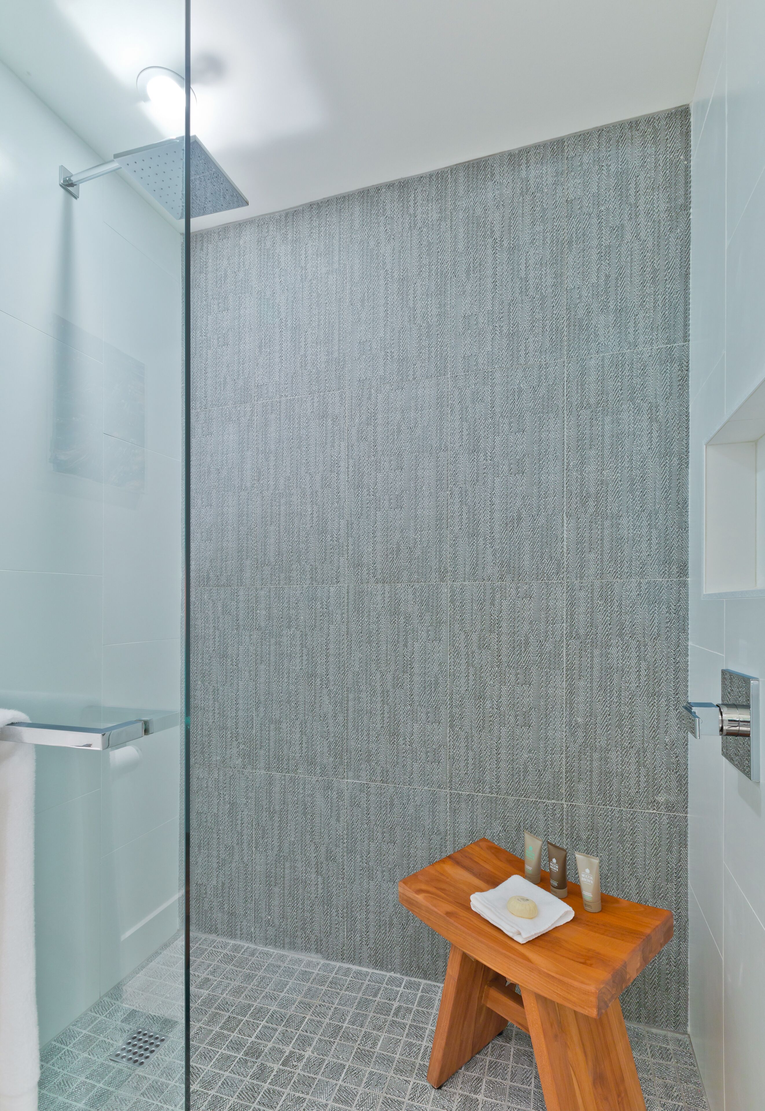 Ein modernes Badezimmer mit Duschkabine aus Glas, einem Holzschemel mit Pflegeprodukten und einer Wand in Grau mit Struktur.